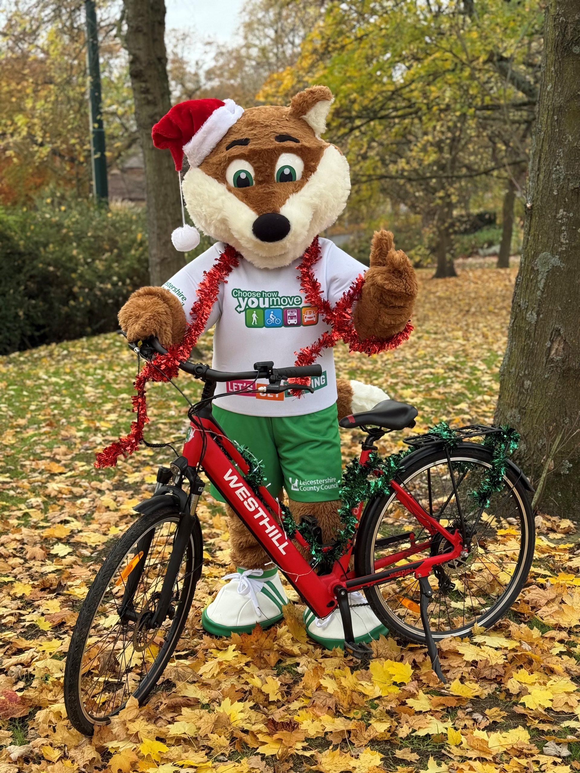 Theo, the Choose How You Move fox mascot, stands behind a bike in a green grassy area with autumn leaves on the floor. Theo is wearing a Santa hat and has tinsel draped around his shoulders to promote the active travel Christmas competition.