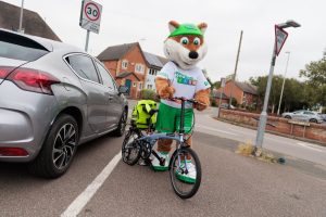 Theo the Fox holding a bicycle with a neon yellow backpack strapped onto the bike - for schools Active Travel Calendar