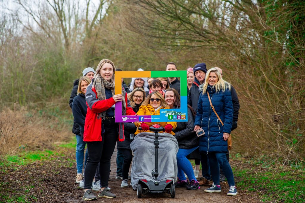 A group people in a wooded area pose with a cardboard frame promoting Choose How You Move