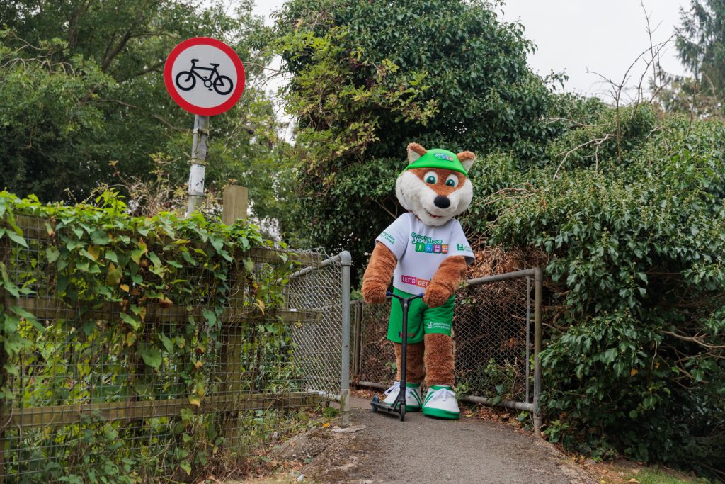 Theo the Fox, the Choose How You Move mascot, rides a scooter across a public footpath, a demonstration of an initiative for the Legacy Grants scheme from Choose How You Move