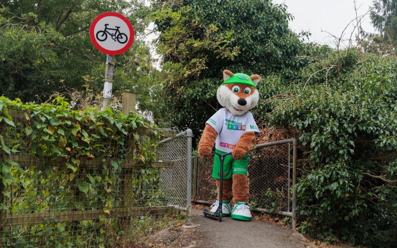 Theo the Fox, the Choose How You Move mascot, rides a scooter across a public footpath, a demonstration of an initiative for the Legacy Grants scheme from Choose How You Move