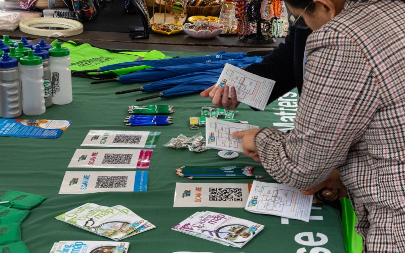 People discussing routes on a Choose How You Move stall, with maps, pencils, bags, umbrellas, and leaflets spread out on the table. For communities and business engagement.