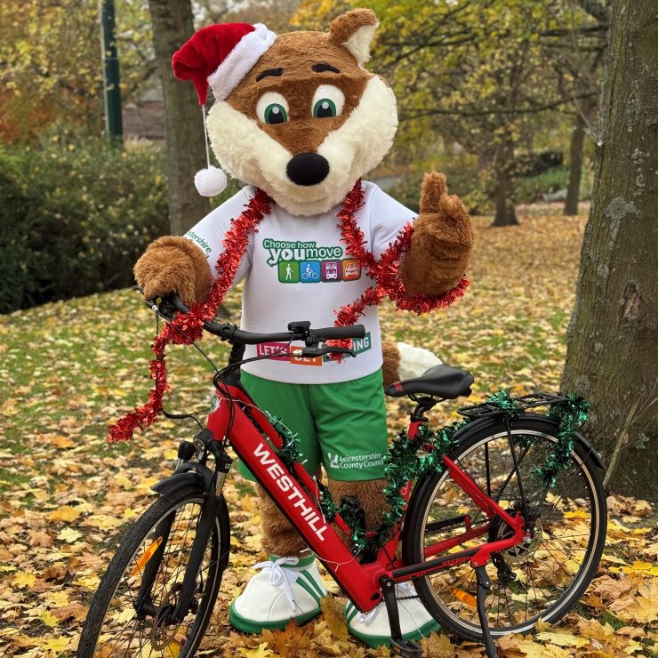 Theo, the Choose How You Move fox mascot, stands behind a bike in a green grassy area with autumn leaves on the floor. Theo is wearing a Santa hat and has tinsel draped around his shoulders.