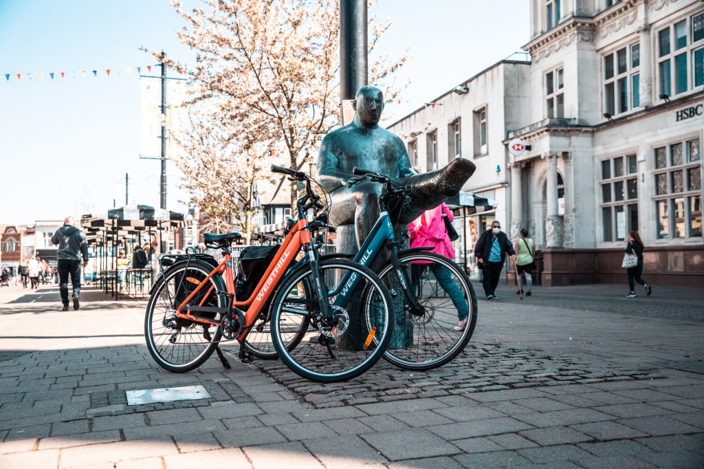 Two electric bikes propped up against a copper statue down a pedestrianised street in Loughborough, Leicestershire.