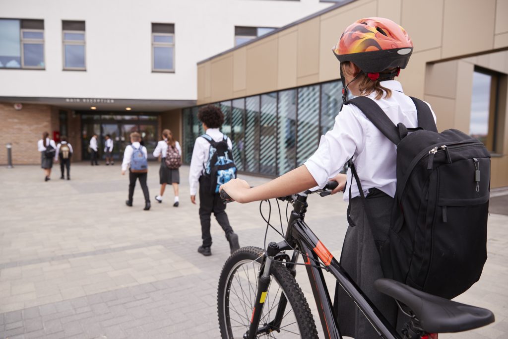 A group of students walking into a British school, with a pupil in the foreground pushing a bike and wearing a helmet
