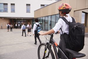 A group of students walking into a British school, with a pupil in the foreground pushing a bike and wearing a helmet