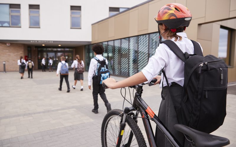 A group of students walking into a British school, with a pupil in the foreground pushing a bike and wearing a helmet