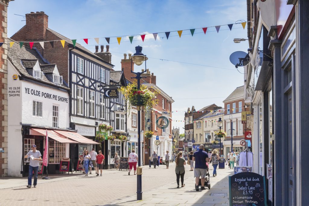 People walking down a high street in Melton Mowbray on a summer’s day, with a decorated lamppost in the centre of the frame and colourful bunting between the buildings. To the left is Melton Mowbray’s famous Ye Olde Porke Pie Shoppe, a white building with a pink awning.