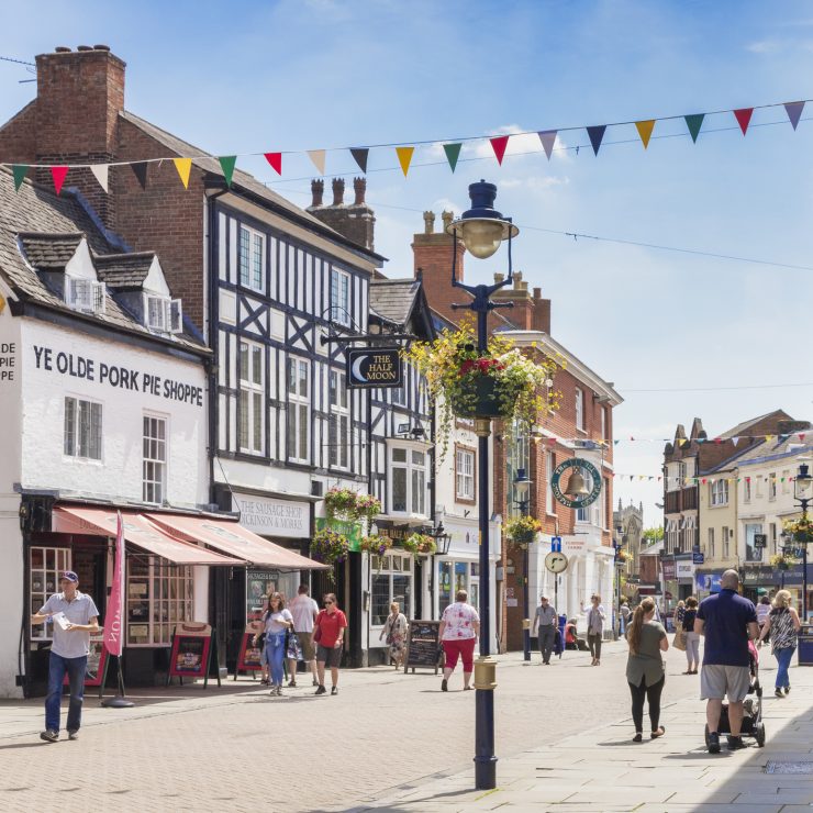 4 July 2019: Melton Mowbray, Leicestershire, UK - People shopping in Nottingham Street on a warm summer day. Ye Olde Pork Pie Shoppe on the left. People walking down a high street in Melton Mowbray on a summer’s day, with a decorated lamppost in the centre of the frame and colourful bunting between the buildings. To the left is Melton Mowbray’s famous Ye Olde Porke Pie Shoppe, a white building with a pink awning.