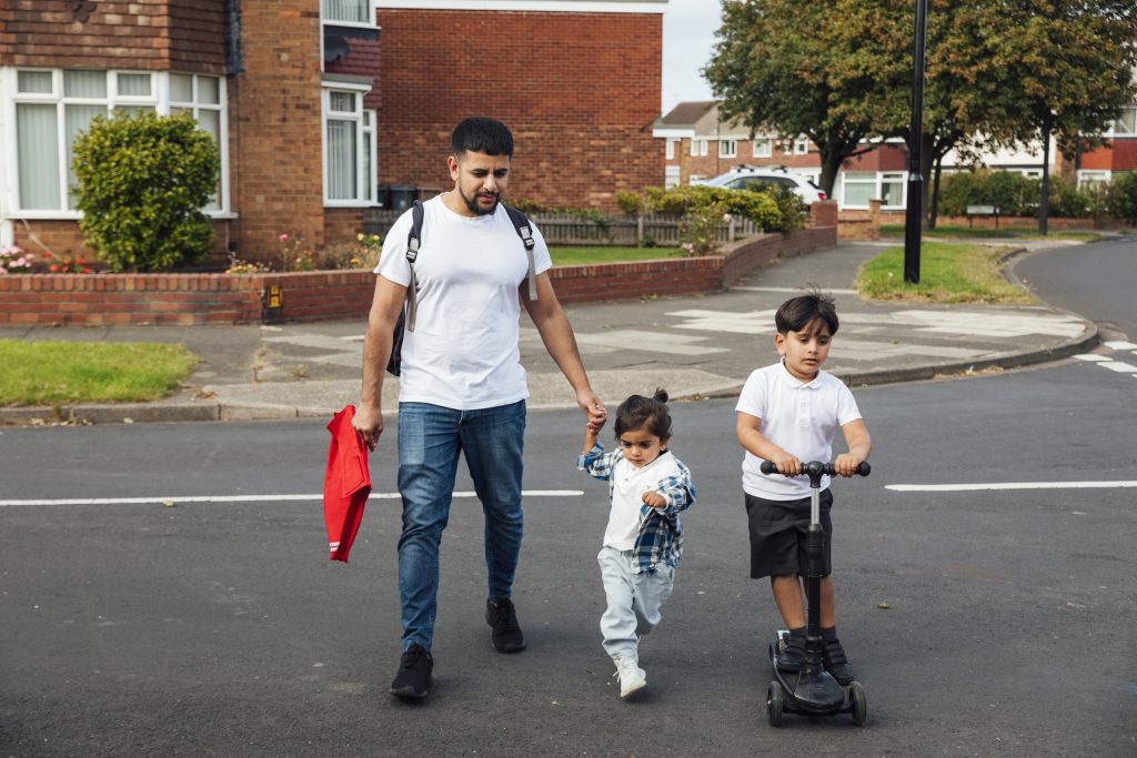 A shot of a father and two young sons crossing a road in a residential area. The older boy is wearing his school uniform and riding on a push scooter. The younger boy is wearing casual clothing and is holding his father's hand while they walk together.