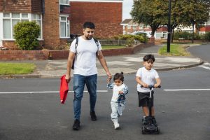 A shot of a father and two young sons crossing a road in a residential area. The older boy is wearing his school uniform and riding on a push scooter. The younger boy is wearing casual clothing and is holding his father's hand while they walk together.