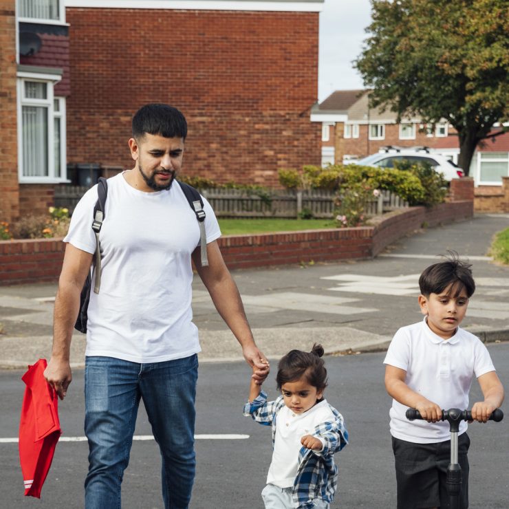 A shot of a father and two young sons crossing a road in a residential area. The older boy is wearing his school uniform and riding on a push scooter. The younger boy is wearing casual clothing and is holding his fathers hand while they walk together. A shot of a father and two young sons crossing a road in a residential area. The older boy is wearing his school uniform and riding on a push scooter. The younger boy is wearing casual clothing and is holding his father's hand while they walk together.