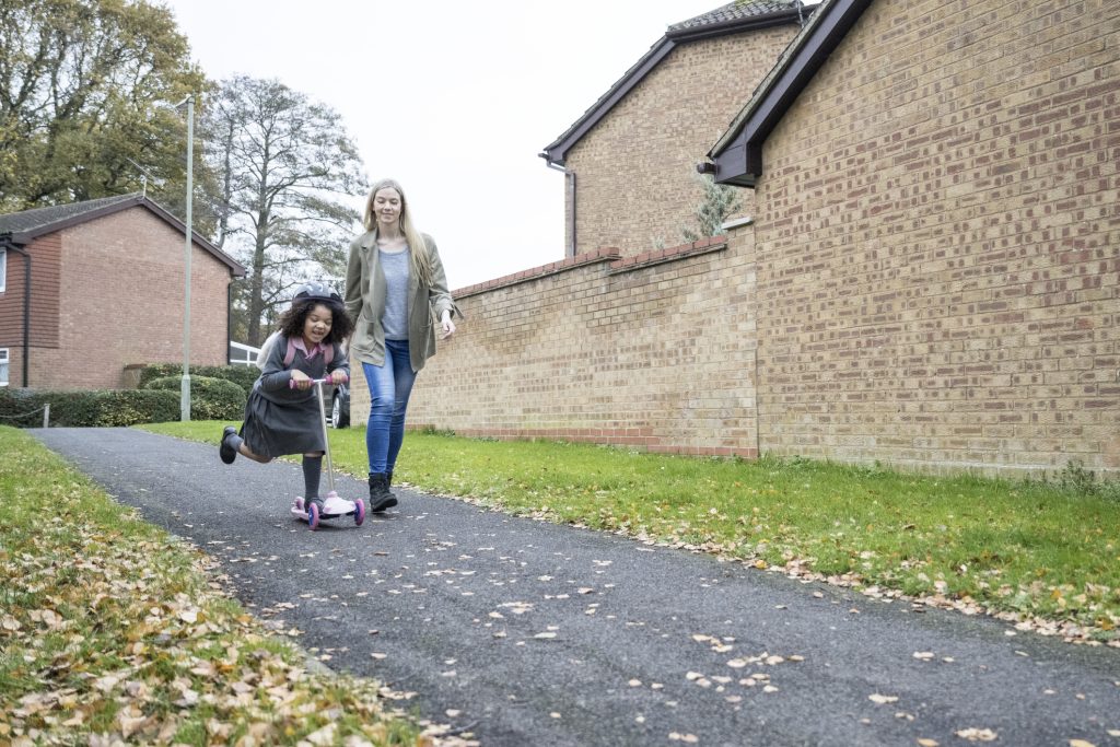A mother in late 30s walking next to 5 year old daughter wearing uniform and riding push scooter on neighbourhood pathway.