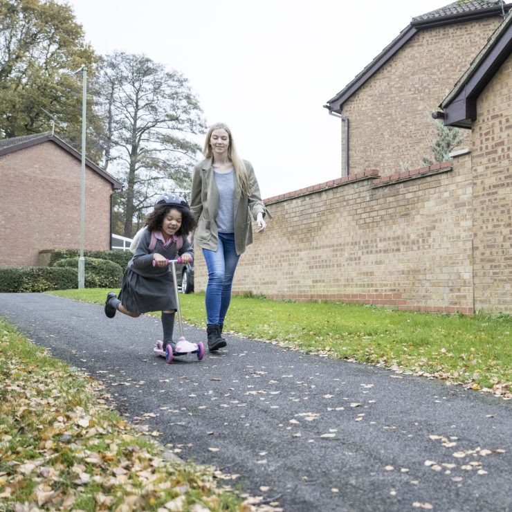 Full length view of Caucasian mother in late 30s walking next to 5 year old daughter wearing uniform and riding push scooter on neighborhood pathway. A mother in late 30s walking next to 5 year old daughter wearing uniform and riding push scooter on neighbourhood pathway.