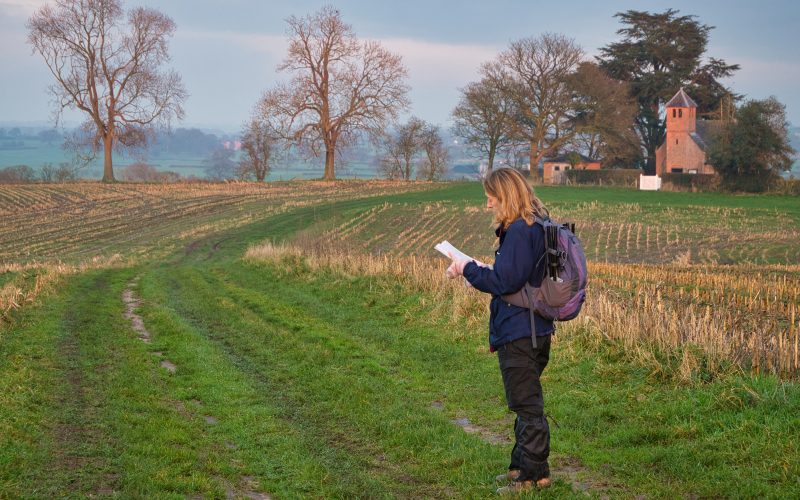 A single woman walker in walking gear on a route through a cut field of crop stubble checks her route with a map. A volunteer for communities work.