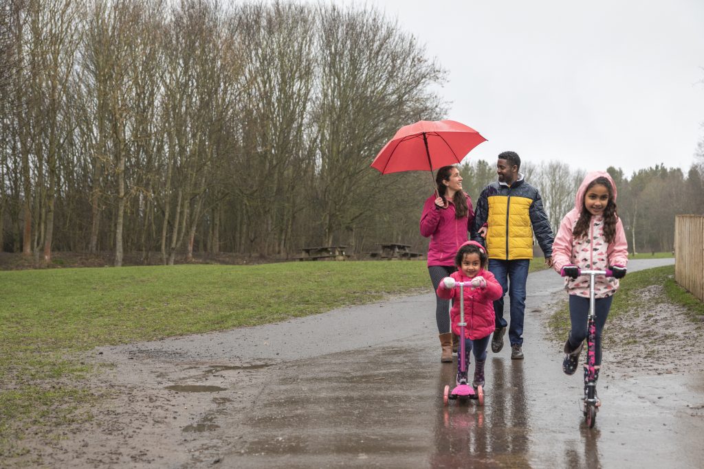 Two young girls riding their push scooters on a wet footpath through a park. Walking behind them, the mother is holding an umbrella above her head to shield her from the rain while talking to her husband.