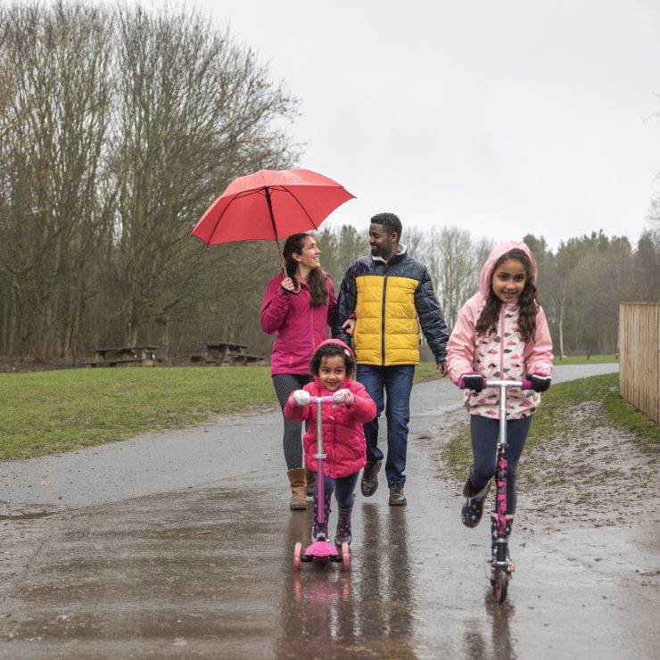 Two young girls riding their push scooters on a wet footpath through Plessey woods, Northumberland. Their parents are walking behind them, the mother is holding an umbrella above her head to shield her from the rain while talking to her husband. They are all enjoying a day out together. Two young girls riding their push scooters on a wet footpath through a park. Walking behind them, the mother is holding an umbrella above her head to shield her from the rain while talking to her husband.