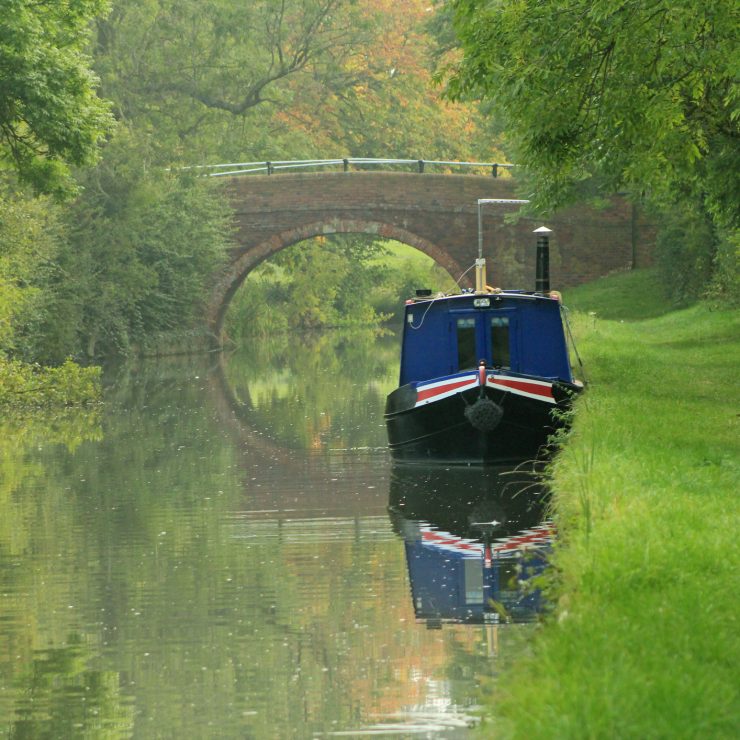 A canal boat moored on a quiet stretch of the Grand Union Canal near Foxton in Leicestershire A blue narrowboat with red detailing moored near Foxton, Leicestershire. Behind the boat is a stone bridge, casting a reflection in the water. For Harborough district.