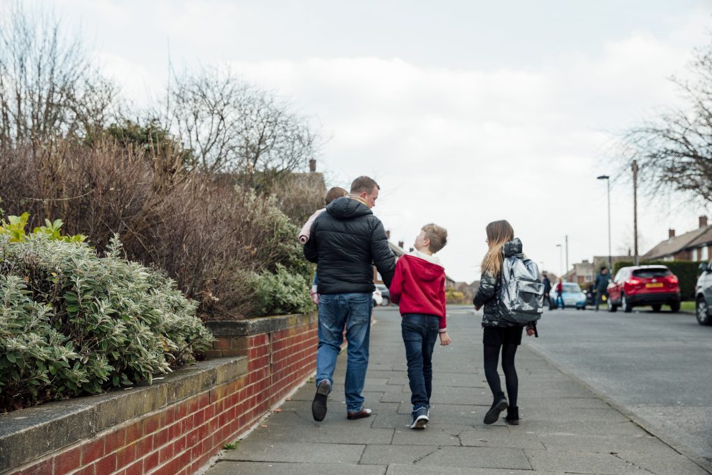 A father holding his toddler and walking with two young children down a neighbourhood path, walking home from school.