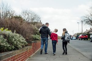 A father holding his toddler and walking with two young children down a neighbourhood path, walking home from school.