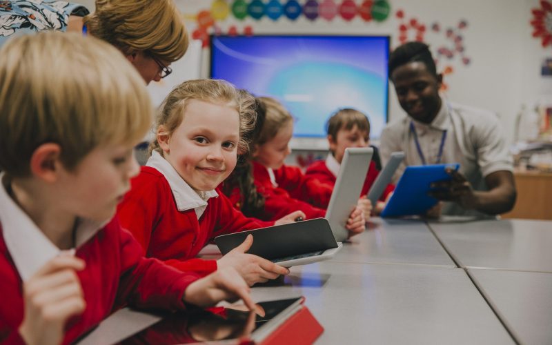 Primary school children are in the classroom, learning about and using digital tablets. One of the girls is smiling at the camera.