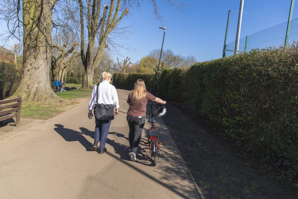 Two people walking down a path in a park: one of them walks with a bike, and one is wearing a laptop bag as if commuting. For cycling, Leicestershire Businesses, business programme.