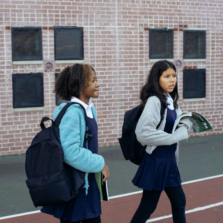 Two schoolgirls in uniform and wearing backpacks walk together across a school playground. The girls are holding a school book on grammar - for schools Active Travel Calendar