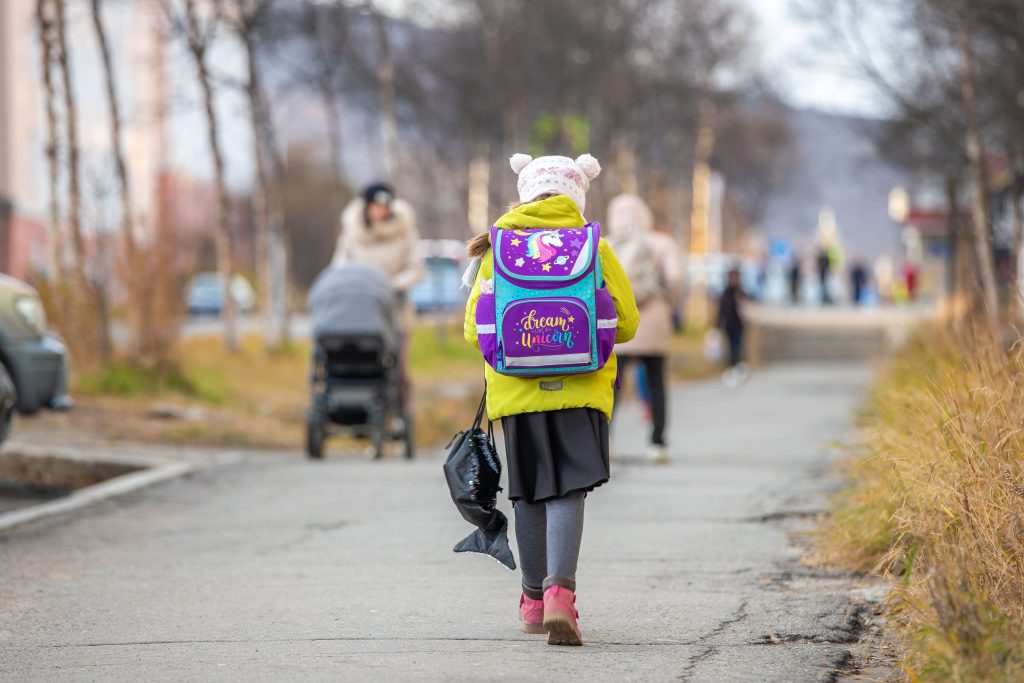 A young child, wearing a yellow coat, a colourful backpack, and a woolly hat with ears, walks down a path. Multiple people walk down the path in the background, including a mother pushing a pram. Walking for schools.