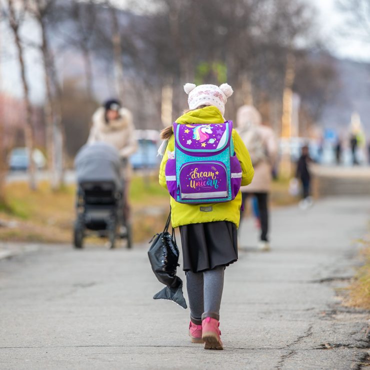 A young child, wearing a yellow coat, a colourful backpack, and a woolly hat with ears, walks down a path. Multiple people walk down the path in the background, including a mother pushing a pram. Walking for schools.