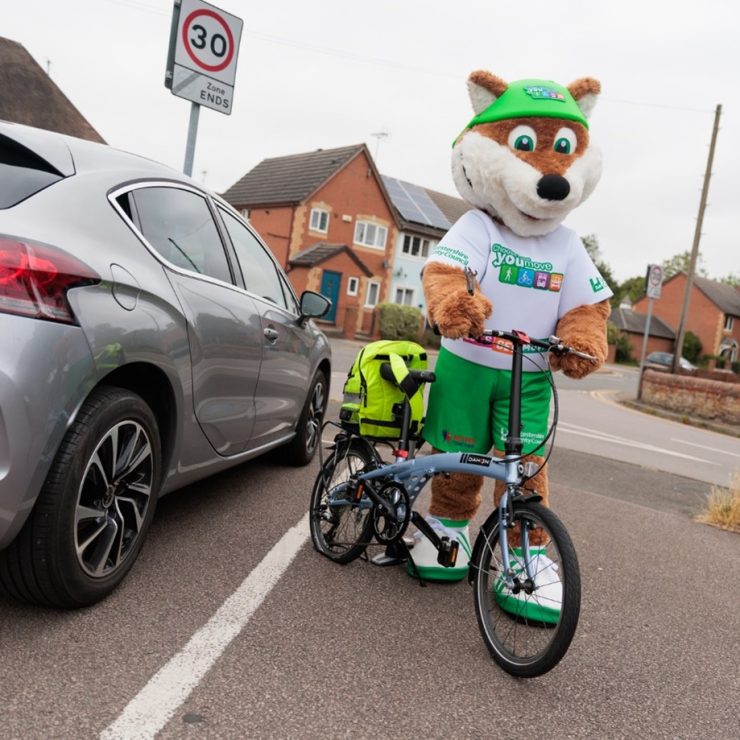 Theo the Fox holding a bicycle with a neon yellow backpack strapped onto the bike - for schools Active Travel Calendar