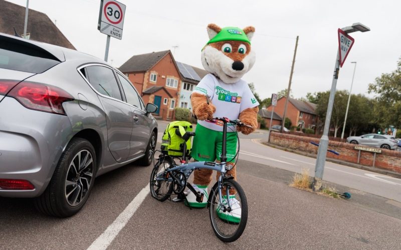 Theo the Fox holding a bicycle with a neon yellow backpack strapped onto the bike - for schools Active Travel Calendar