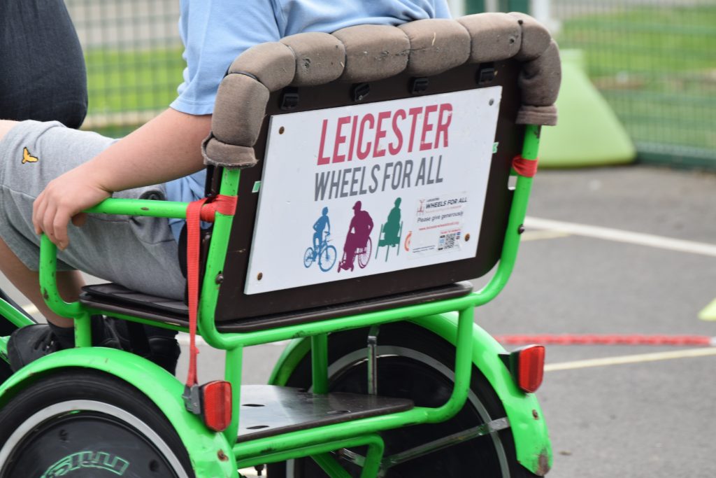 a child in a blue shirt sits in a neon green adapted cycle. The sign on the seat displays the logo for Leicester Wheels For All. For accessible transport and promoting adaptive bikes.