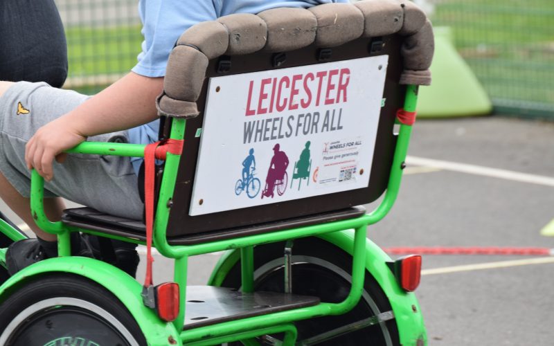 a child in a blue shirt sits in a neon green adapted cycle. The sign on the seat displays the logo for Leicester Wheels For All. For accessible transport and promoting adaptive bikes.