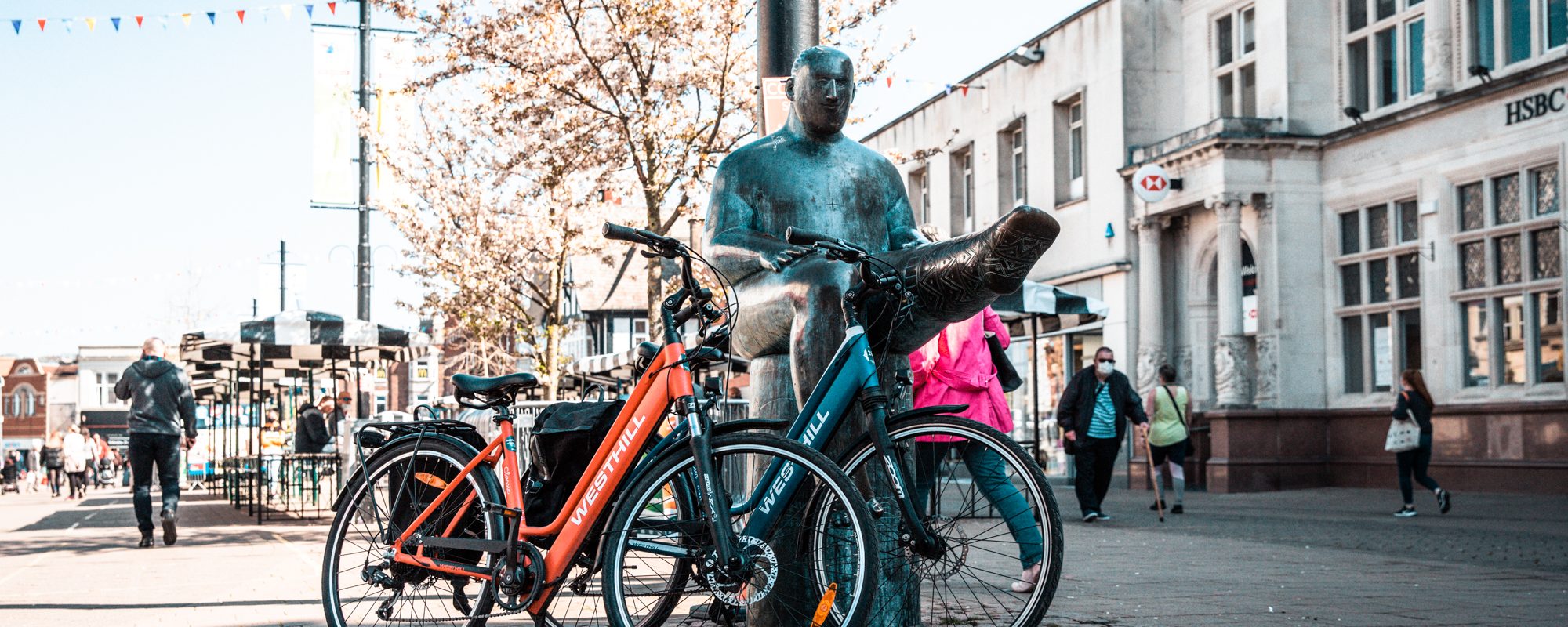 Two electric bikes propped up against a copper statue down a pedestrianised street in Loughborough, Leicestershire.