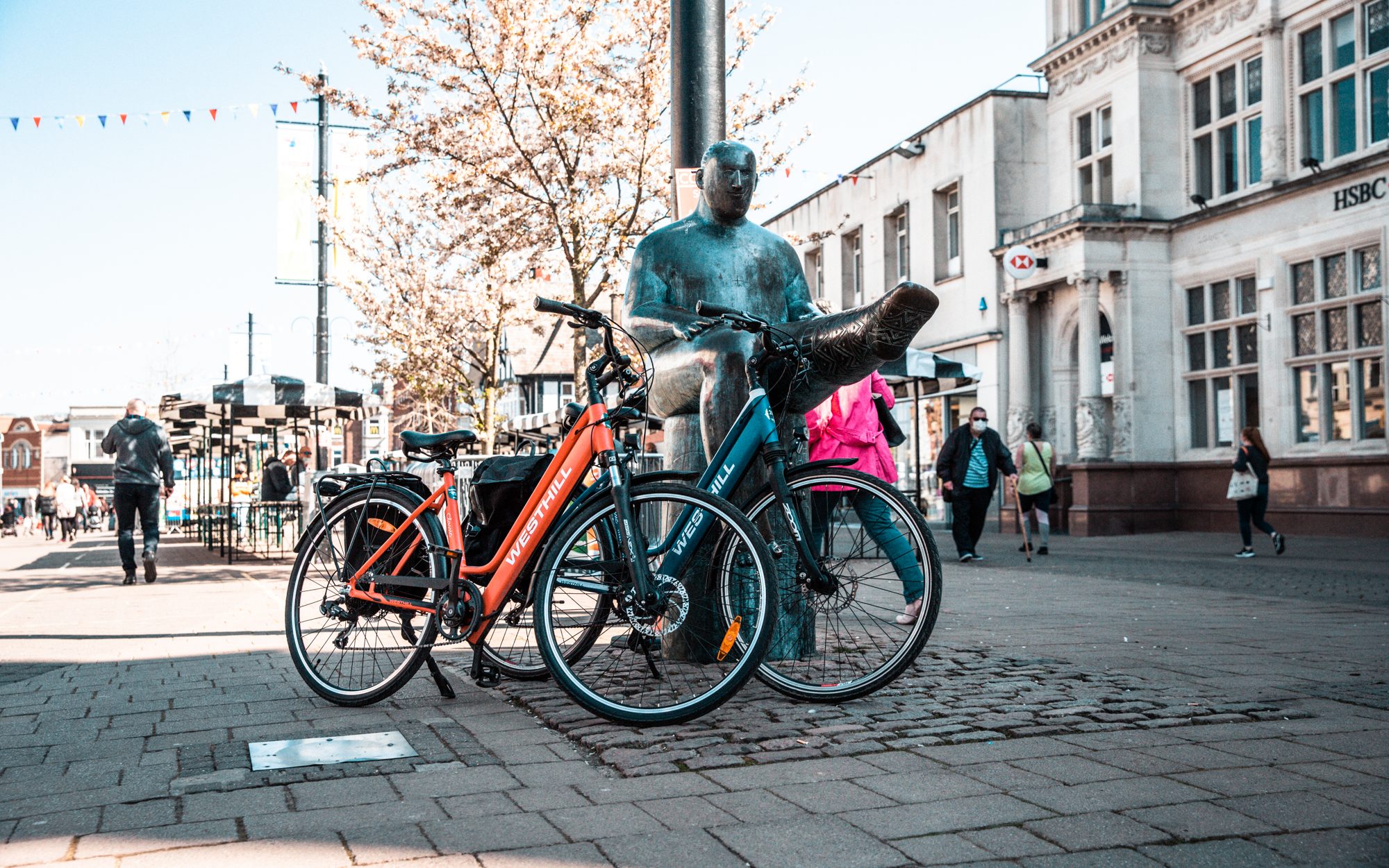 Two electric bikes propped up against a copper statue down a pedestrianised street in Loughborough, Leicestershire.