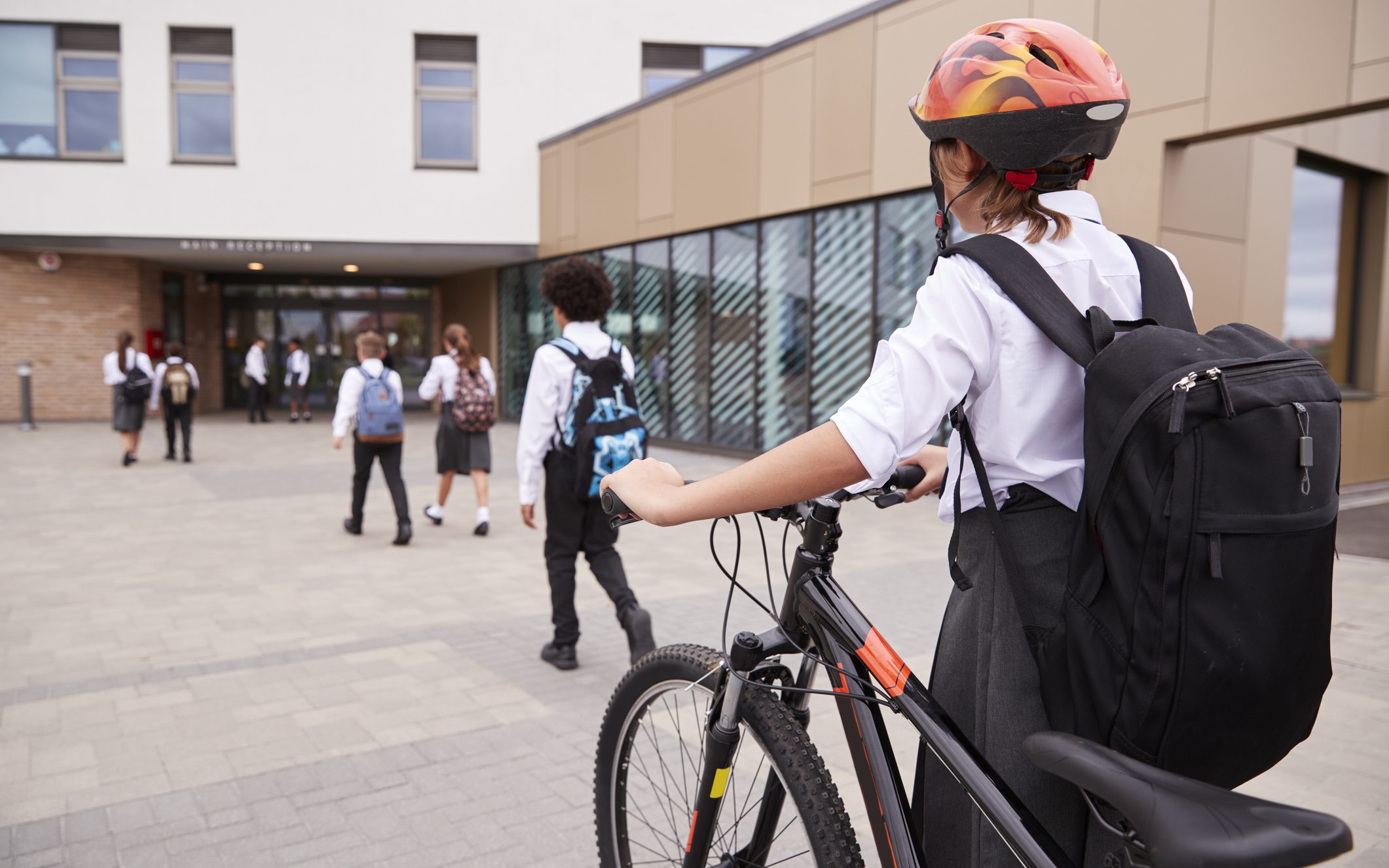 A group of students walking into a British school, with a pupil in the foreground pushing a bike and wearing a helmet