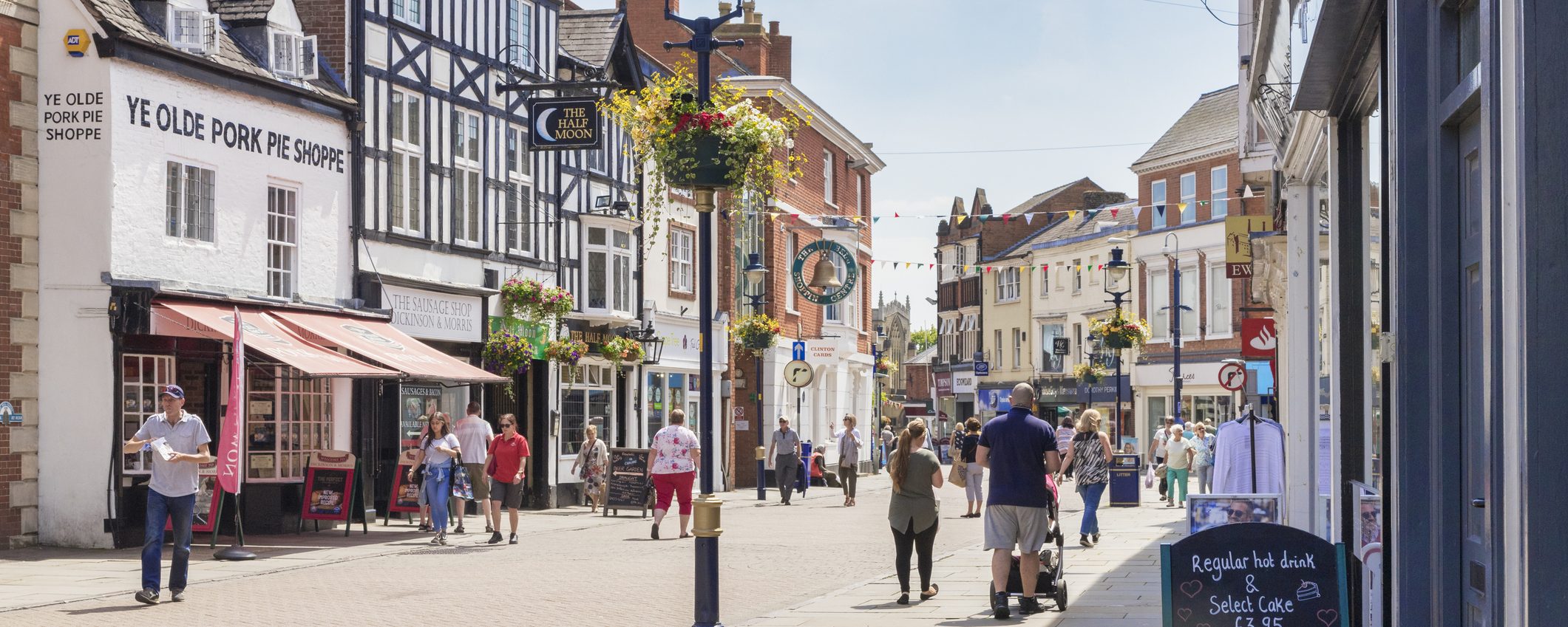 People walking down a high street in Melton Mowbray on a summer’s day, with a decorated lamppost in the centre of the frame and colourful bunting between the buildings. To the left is Melton Mowbray’s famous Ye Olde Porke Pie Shoppe, a white building with a pink awning.