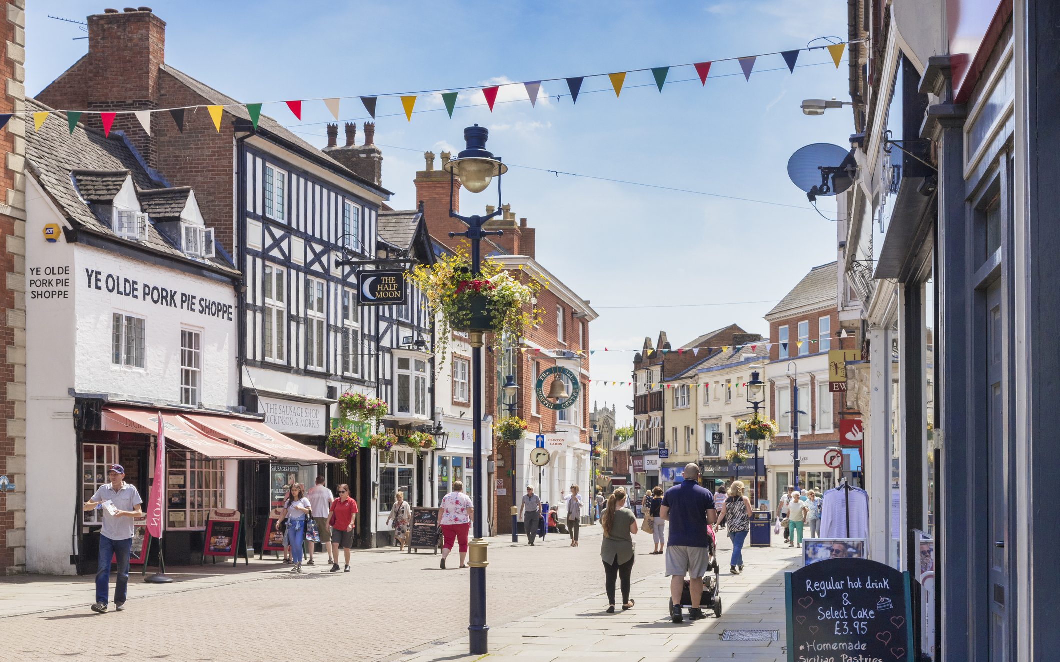 People walking down a high street in Melton Mowbray on a summer’s day, with a decorated lamppost in the centre of the frame and colourful bunting between the buildings. To the left is Melton Mowbray’s famous Ye Olde Porke Pie Shoppe, a white building with a pink awning.