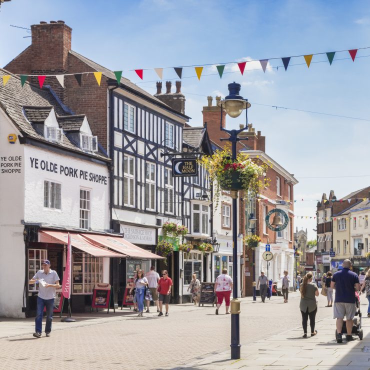 People walking down a high street in Melton Mowbray on a summer’s day, with a decorated lamppost in the centre of the frame and colourful bunting between the buildings. To the left is Melton Mowbray’s famous Ye Olde Porke Pie Shoppe, a white building with a pink awning.