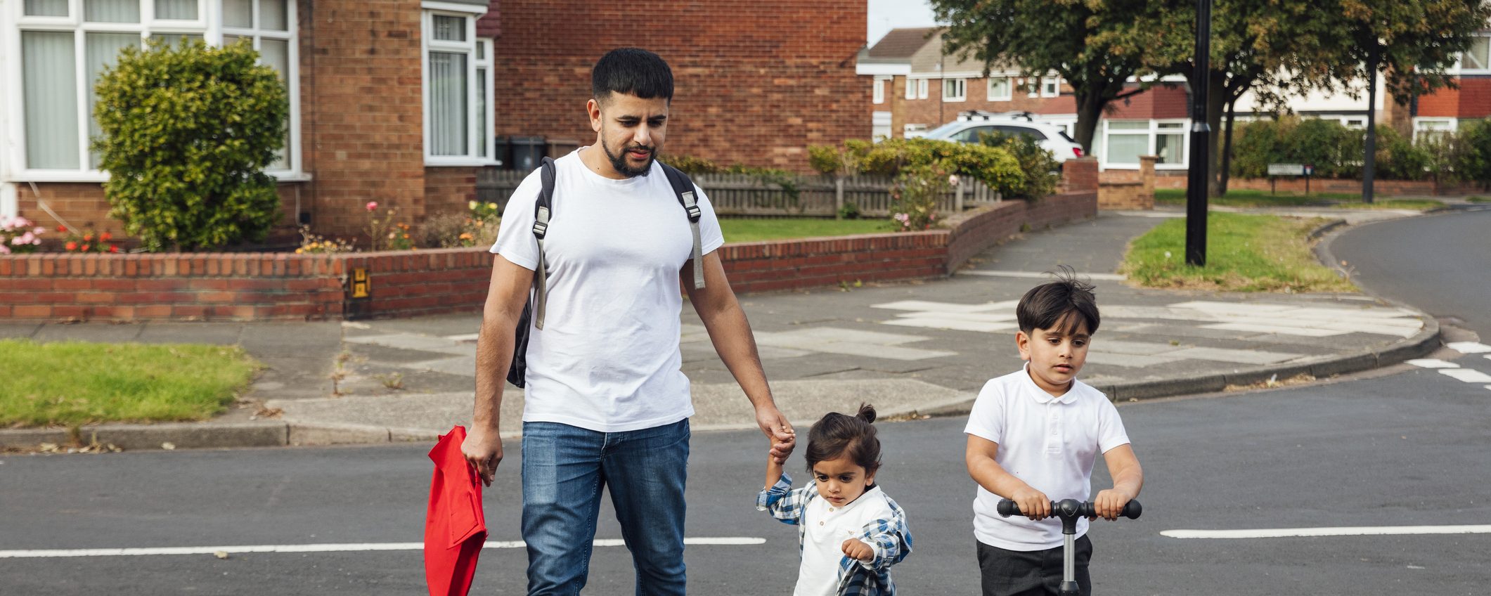 A shot of a father and two young sons crossing a road in a residential area. The older boy is wearing his school uniform and riding on a push scooter. The younger boy is wearing casual clothing and is holding his father's hand while they walk together.