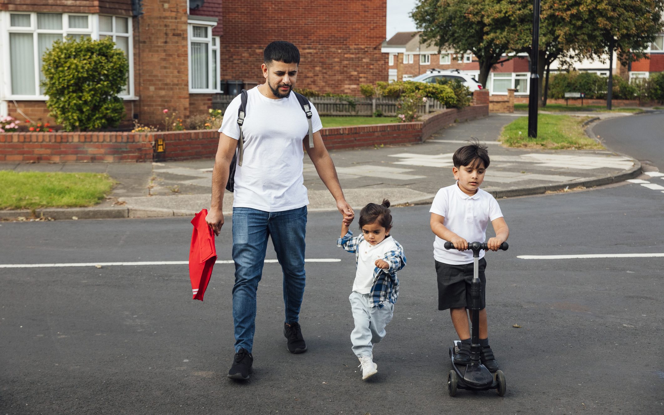 A shot of a father and two young sons crossing a road in a residential area. The older boy is wearing his school uniform and riding on a push scooter. The younger boy is wearing casual clothing and is holding his father's hand while they walk together.