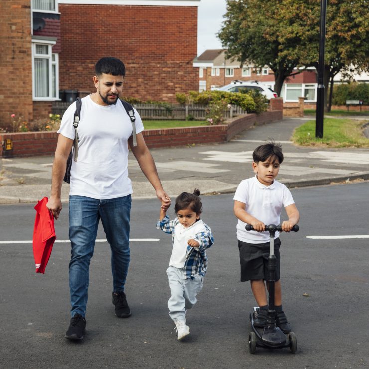 A shot of a father and two young sons crossing a road in a residential area. The older boy is wearing his school uniform and riding on a push scooter. The younger boy is wearing casual clothing and is holding his father's hand while they walk together.