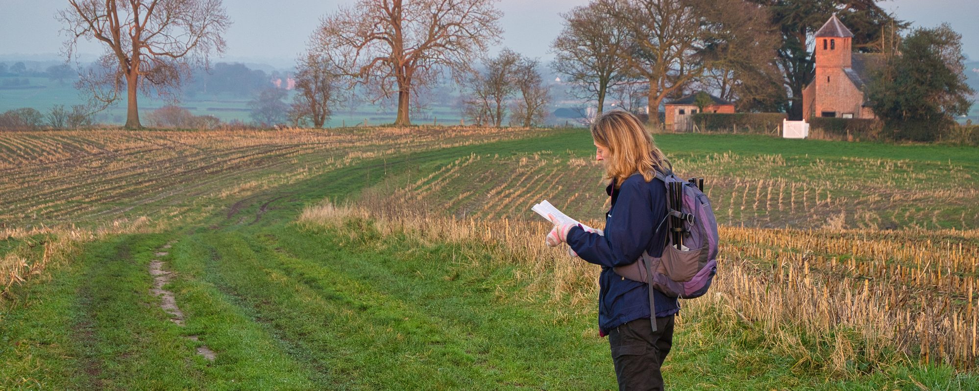 A single woman walker in walking gear on a route through a cut field of crop stubble checks her route with a map.