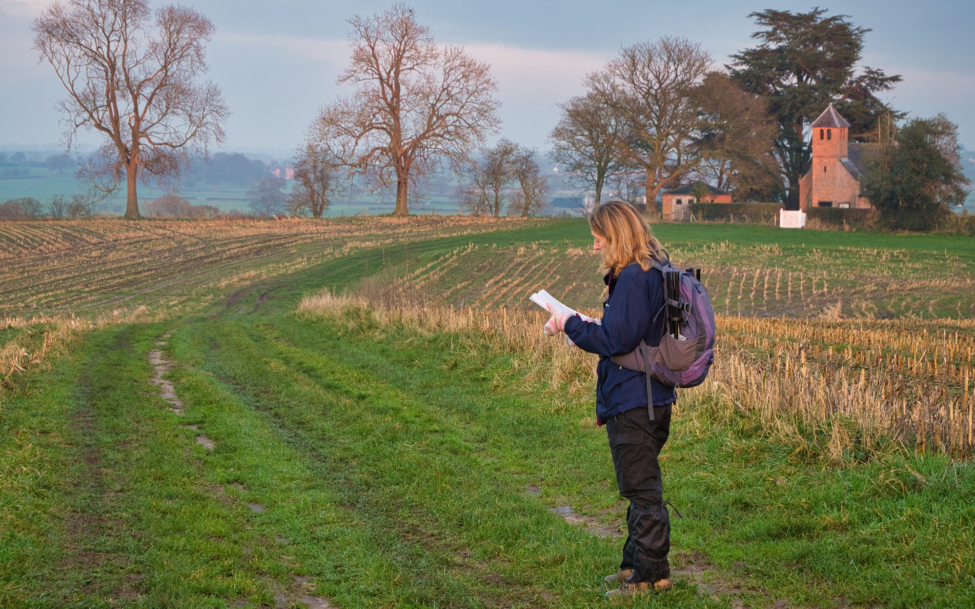 A single woman walker in walking gear on a route through a cut field of crop stubble checks her route with a map.