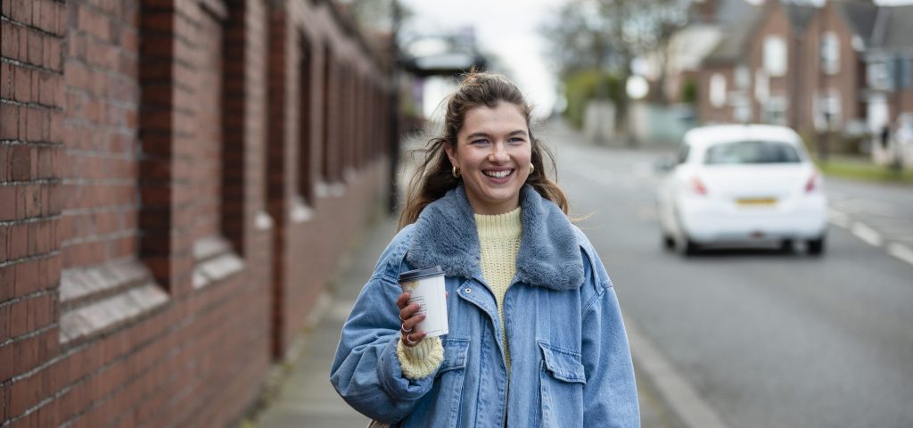 A woman in a blue jacket holding a coffee and walking to work. For commuting, Leicestershire Businesses, business programme.