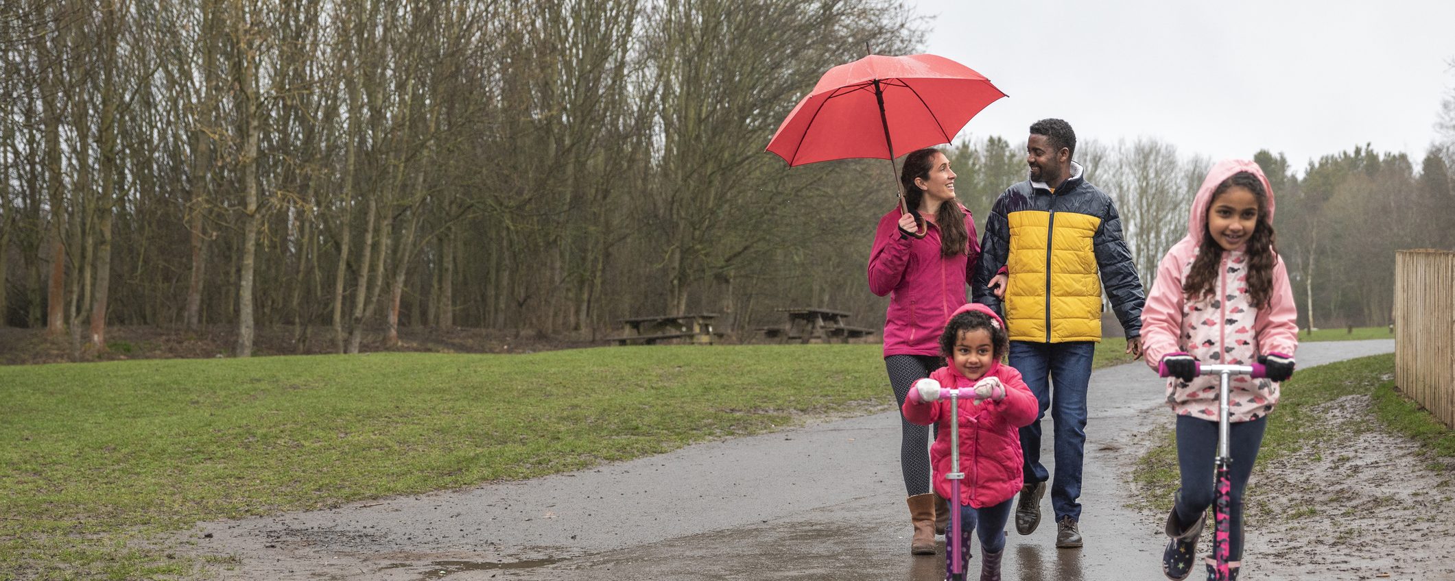 Two young girls riding their push scooters on a wet footpath through a park. Walking behind them, the mother is holding an umbrella above her head to shield her from the rain while talking to her husband.