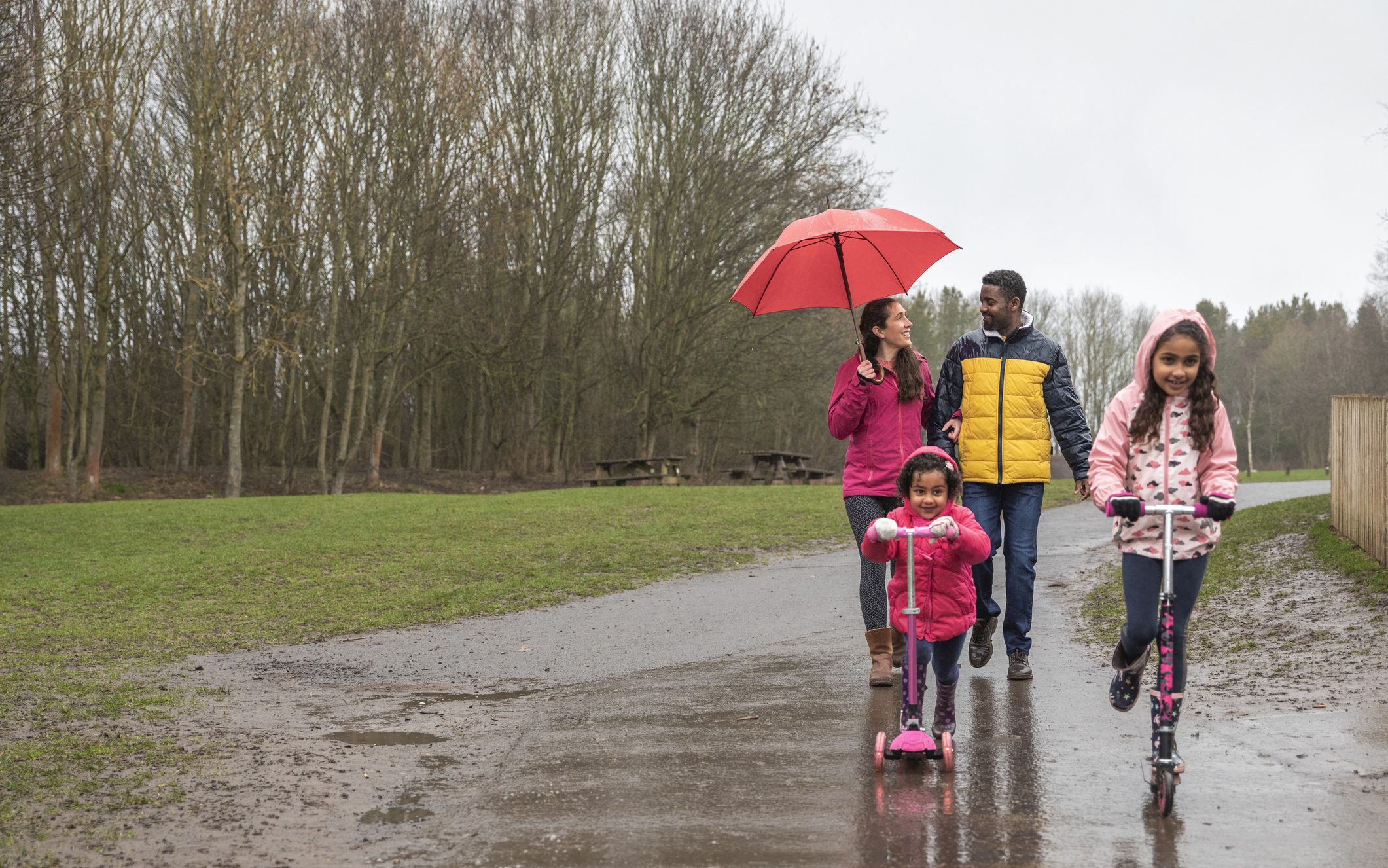 Two young girls riding their push scooters on a wet footpath through a park. Walking behind them, the mother is holding an umbrella above her head to shield her from the rain while talking to her husband.