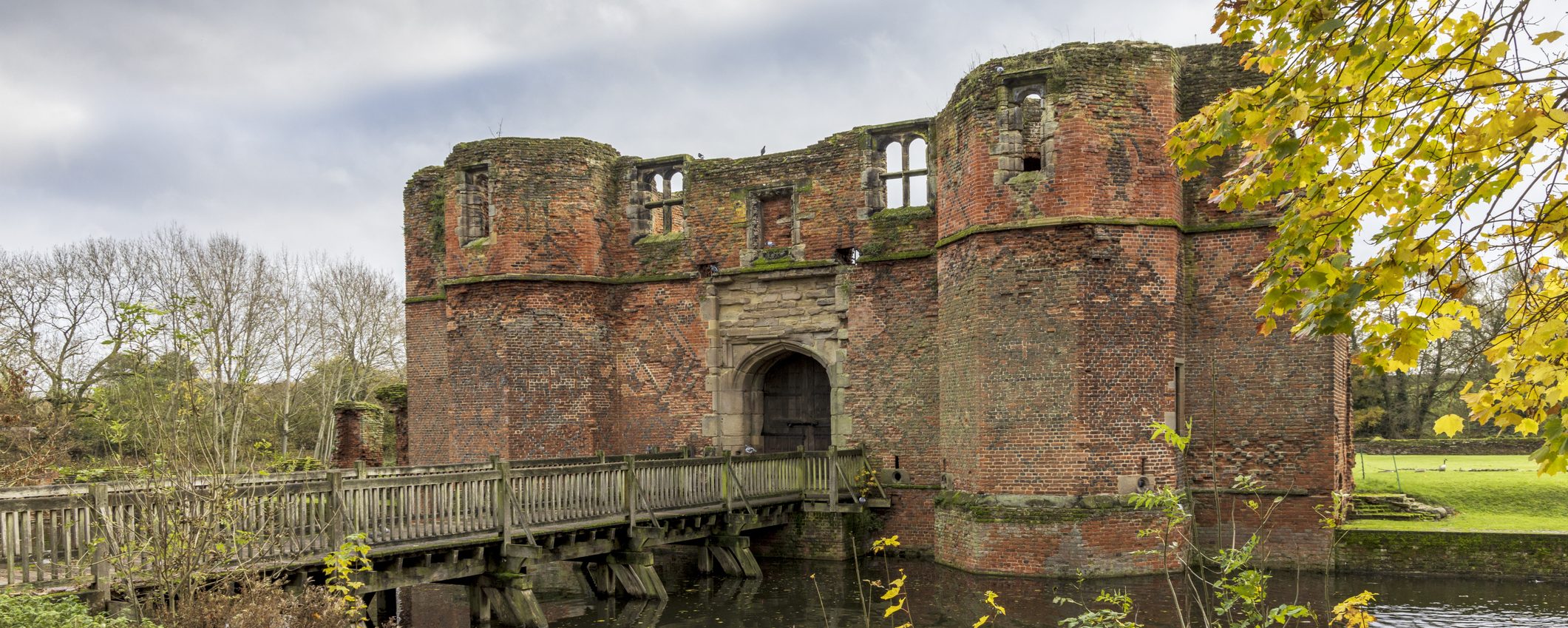A bridge on a moat leading up to Kirby Muxloe Castle, a ruined red-brick castle. On the bank, a goose walks amongst yellow leaves. For Blaby district.
