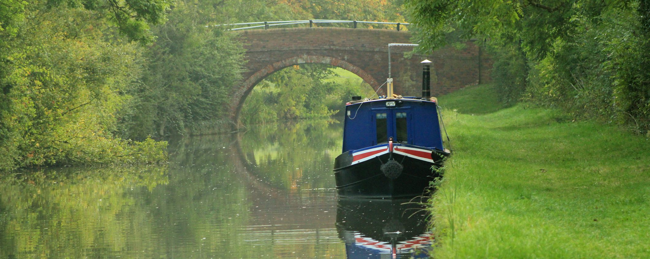 A blue narrowboat with red detailing moored near Foxton, Leicestershire. Behind the boat is a stone bridge, casting a reflection in the water. For Harborough district.