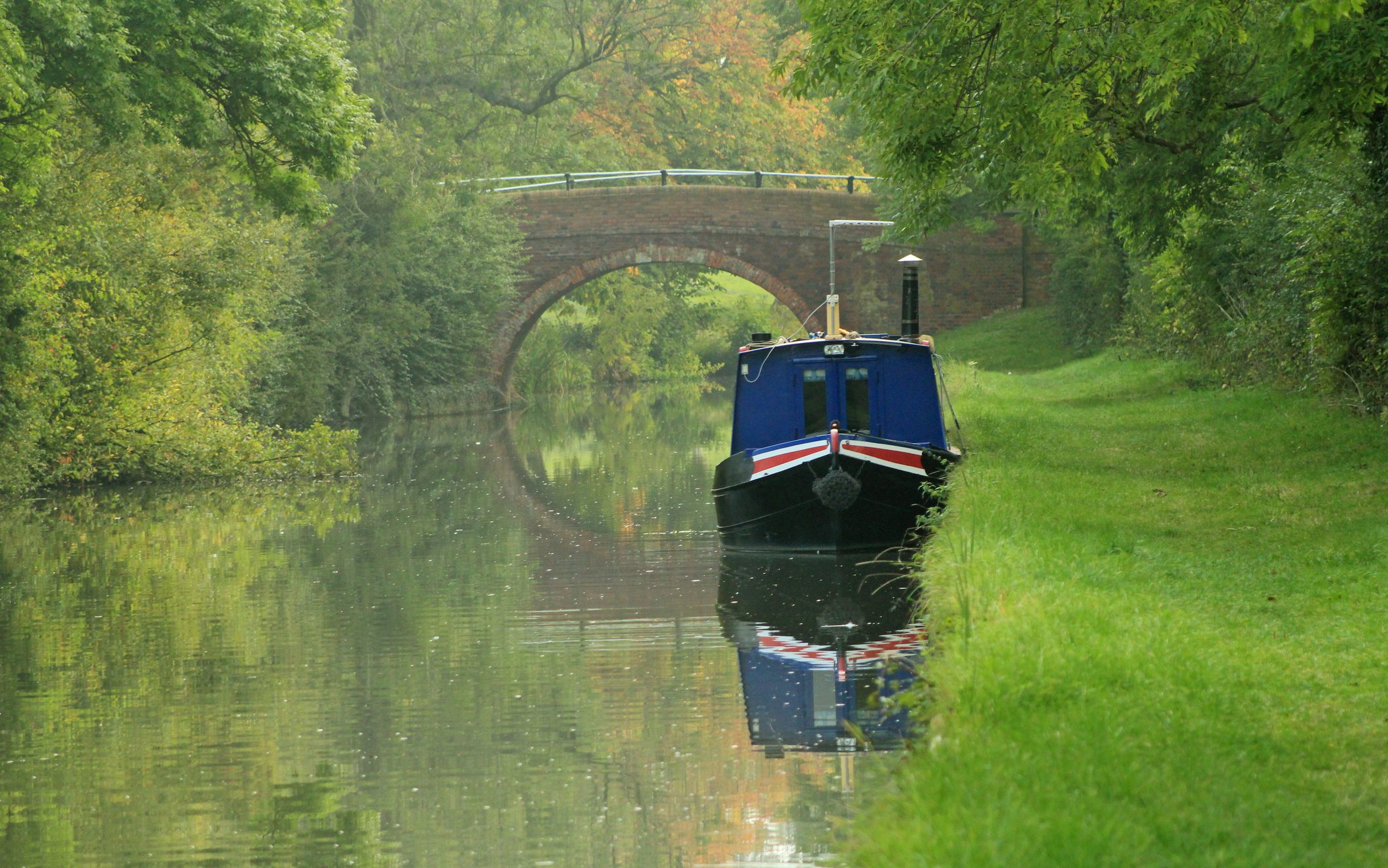 A blue narrowboat with red detailing moored near Foxton, Leicestershire. Behind the boat is a stone bridge, casting a reflection in the water. For Harborough district.